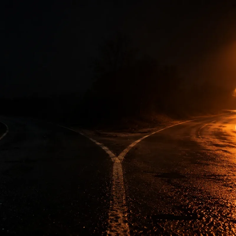 A night road splitting in two, one side lit by warm street lamps and the other disappearing into darkness
